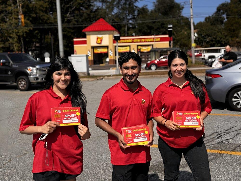 Three street team members in red polos hold grand opening coupons outside a new auto service shop