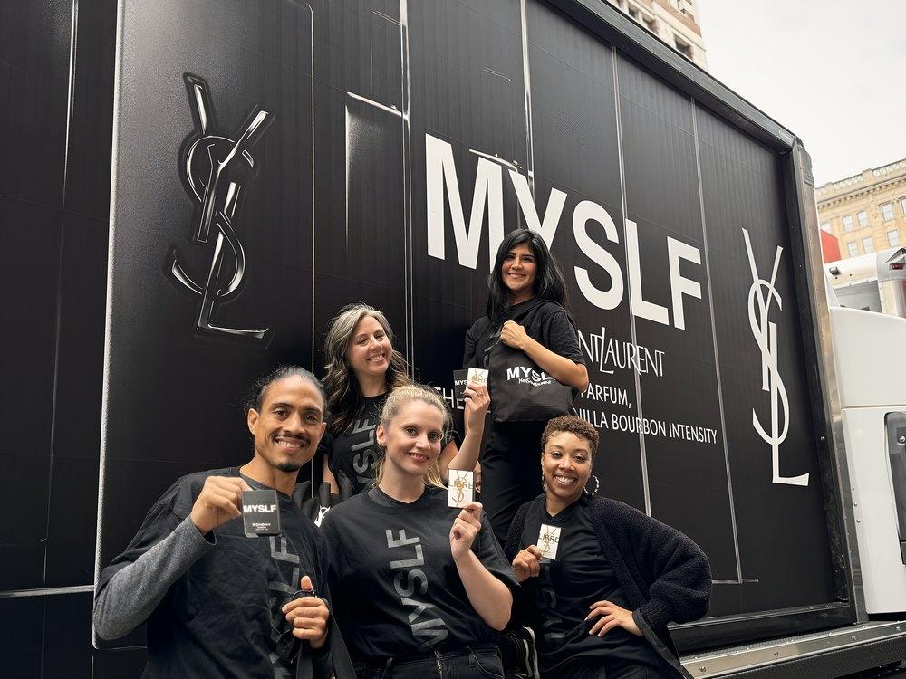 Five brand ambassadors in black tees pose with fragrance samples beside a black branded truck in the city