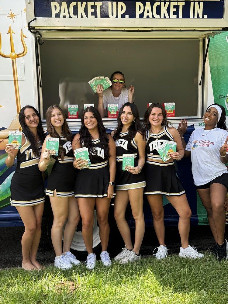 College cheer team and two brand staff hand out snack packets from a food truck window on a campus lawn